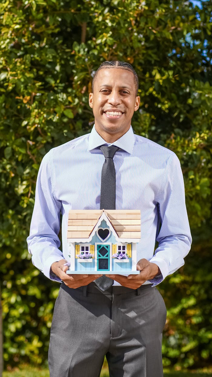 Services Smiling real estate agent holding a model house in an outdoor setting.